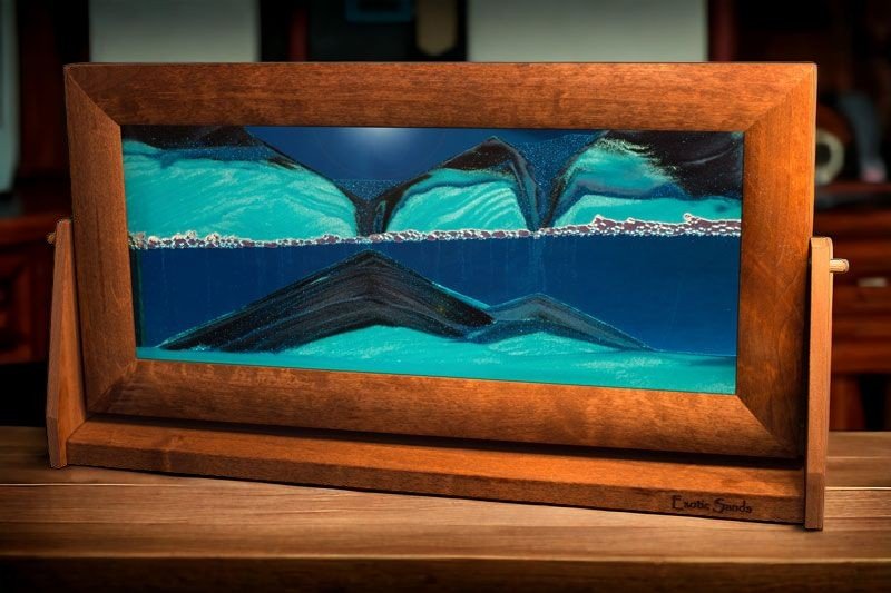 Extra-large Sandscape Sand Picture featuring flowing black and white sands suspended in vibrant blue liquid. Encased in an alder wood frame with a swivel stand, this mesmerizing, ever-changing sand art adds a dynamic touch to any home or office.